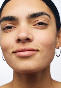 Gros plan sur un visage avec une peau lisse, un maquillage naturel et des taches de rousseur subtiles. Présente des boucles d'oreilles en argent et des cheveux lisses et foncés.
