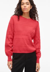 Vrouw met krullend haar, gekleed in een gerstekend rood gebreide trui en zwarte broek, staand tegen een effen witte achtergrond.