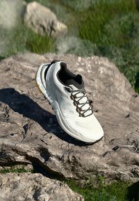 Weißer Laufschuh mit strukturiertem Obermaterial, schwarzem Innenbereich und gelben Akzenten. Besitzt atmungsaktives Material und flache Schnürsenkel. Auf einem Stein platziert.