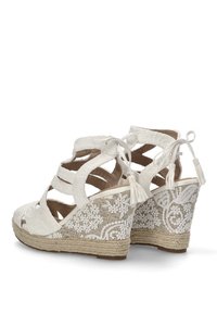 Sandalias de cuña blancas con una parte superior de tela texturizada, diseño floral bordado en el talón y borlas decorativas en las cintas del tobillo.