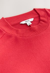 Rode trui met een geribde kraag en een textuurstof. Label zegt 'NEXT.' Close-up van het ontwerp van de halslijn en het materiaal.