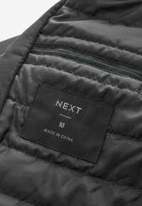 Schwarze, gepolsterte Jacke mit gestepptem Innenfutter. Enthält ein Etikett mit der Aufschrift "NEXT", Größe "M" und "MADE IN CHINA". Keine weiteren Details.