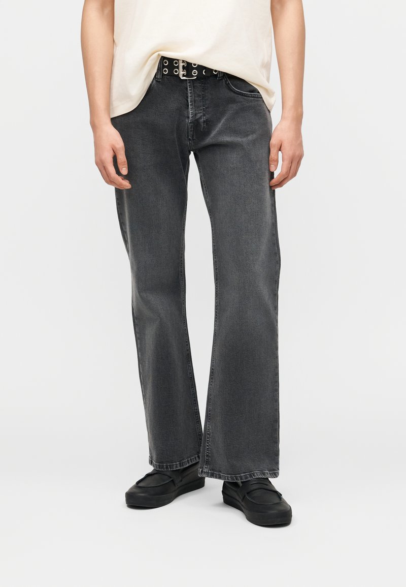 Άτομο που φοράει φαρδιά ξεθωριασμένα μαύρα τζιν, μαύρη ζώνη με ασημί μεταλλικά τρύπες, μαύρα slip-on παπούτσια και κρεμ μπλούζα μπλουζάκι που έχει βάλει μέσα στο παντελόνι.