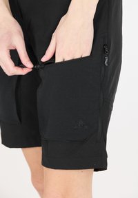 Mustat shortsit, joissa on vetoketjulliset sivutaskut, valmistettu kevyestä kankaasta, jossa on sileä pinta ja pieni logo helman kohdalla.