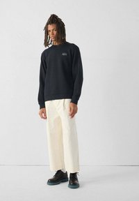 Sudadera negra con pequeño logo, pantalones anchos cremosos y zapatos negros con suela color teal. El modelo está de pie contra un fondo sencillo.