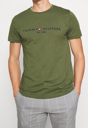 Mand iført en olivengrøn Tommy Hilfiger t-shirt med logo og tekst "EST. 1985" kombineret med lysegrå ternede bukser med hænderne i lommerne.