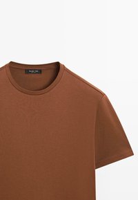 T-shirt en coton marron avec un col rond classique et des manches courtes. Texture douce, finition lisse, et une étiquette de marque discrète sur le col.