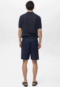 Polo shirt in maglina navy a maniche corte, abbinato a pantaloni navy. Entrambi i capi presentano una finitura testurizzata. Le calzature sono sandali neri.