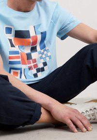 Light blue cotton t-shirt featuring a geometric, multicolored abstract print in reds, blues, and whites. Round neckline, short sleeves.