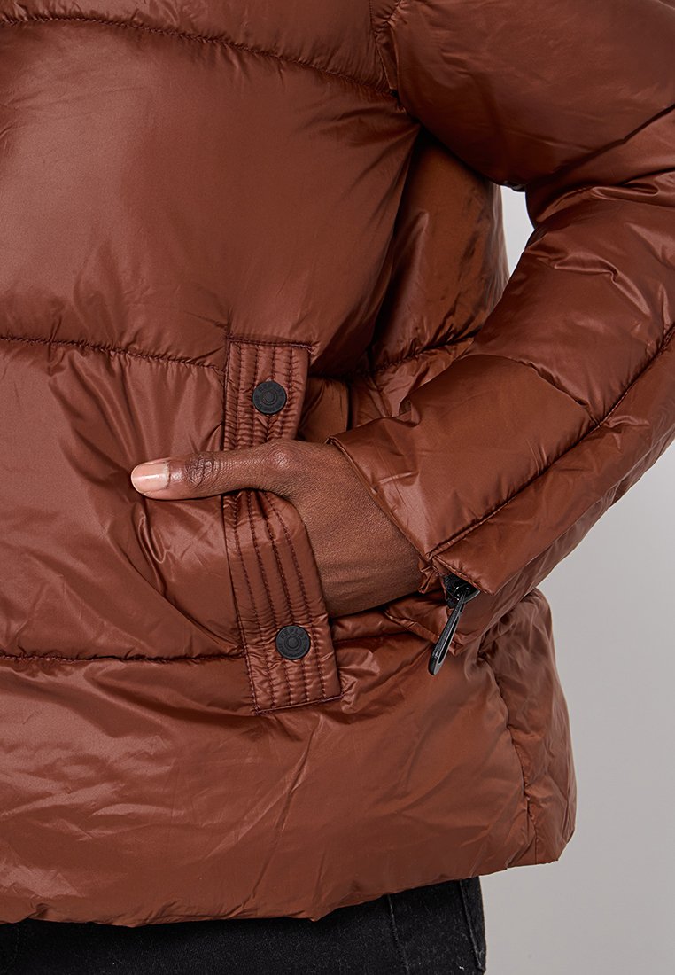 Giacca a vento marrone con una texture liscia e lucida. Presenta ampie cuciture, bottoni automatici e tasche con zip sui lati.