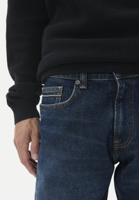 Tamsiai mėlyni džinsai su klasika, turintys auksinę siūlę, priekinį kišenę su grioveliu ir tekstūruotą juodą megztinį viršuje.