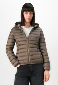 Vrouw met steil donker haar in een lichtbruine gewatteerde jas met capuchon en zwarte jeans, staand tegen een witte achtergrond.