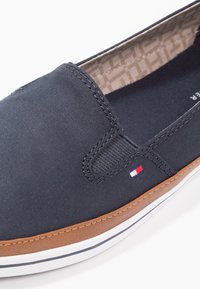 Navy canvas slip-on shoe with brown faux leather sole, white horizontal stripes, and small logo patch in red, white, and blue on the side.