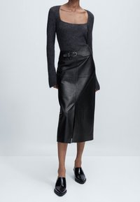 Jupe midi en cuir noir avec une fente latérale, dotée d'un détail boucle de ceinture. Associée à un haut en maille à manches longues gris foncé et des mules noires brillantes.