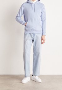 Světle modrá mikina s kapucí a přední kapsou, doplněná o světlé džínové kalhoty s rovnými nohavicemi a bílé tenisky s černými detaily. Jednoduchý design.