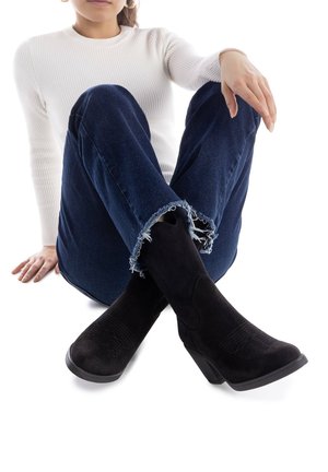Botas altas de ante negro con tacón cuadrado, que presentan sutiles detalles de textura y una punta redondeada, combinadas con jeans de mezclilla oscura y un suéter blanco.