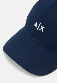 Boné azul-escuro feito de tecido com uma textura suave, apresentando um logotipo bordado em branco "A/X" na frente e uma aba estruturada.