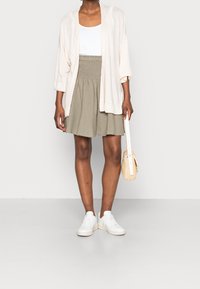 Cardigan léger rose pâle ouvert, débardeur blanc, jupe vert olive avec taille froncée, baskets blanches, tenant un petit sac tissé. Tenue simple et décontractée.