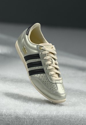 Basket en cuir gris clair avec des rayures noires, détails perforés, lacets de couleur crème et accents dorés pour le logo. Semelle en caoutchouc pour l'adhérence.