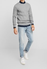 Szary sweter w splot kabelkowy nałożony na granatową bluzkę z kołnierzykiem, w połączeniu z jasnoniebieskimi dżinsami i białymi trampkami. Całość charakteryzuje się teksturowanymi wzorami.