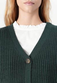 Kvinne iført en mørkegrønn ribbestrikket cardigan med en enkelt knapp over en hvit rynket kragebluse, vist fra hake til bryst.