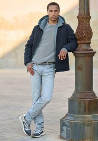 Men's outfit featuring a navy jacket with a grey hood, ribbed grey sweater, light blue jeans, and grey-and-white sneakers. Standing by a streetlamp.