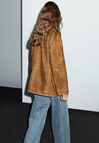 Giacca in suede marrone con una vestibilità rilassata, cuciture visibili e maniche larghe, abbinata a jeans a gamba larga in lavaggio chiaro su uno sfondo minimalista.