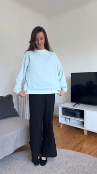 Γυναίκα μέσα στο σπίτι με ανοιχτό μπλε φούτερ και μαύρο παντελόνι φαρδιάς γραμμής, στέκεται σε στρογγυλό μπεζ χαλί κοντά σε καναπέ και έπιπλο τηλεόρασης.