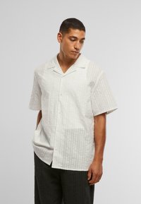 Camicetta bianca a maniche corte con un motivo a righe testurizzato, chiusura con bottoni e colletto classico. Indossata con pantaloni scuri e con motivo.