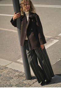 Tume topeltkarvaga mantel kuldsete nööpidega, mille all on valge kõrge kaelusega kampsun, kombineeritud laia triibuliste pükstega ja mustade pahkluu saapadega.