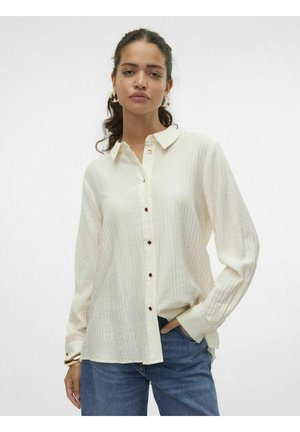 Camisa de botones color crema con cuello, tejido texturizado, botones en tono dorado y mangas largas, combinada con vaqueros azules.
