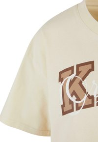 Beige cotton t-shirt featuring a large brown and white graphic design with the letter "K". Short sleeves and a crew neck.