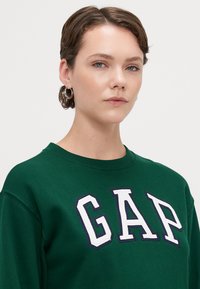 Grønn hettegenser med "GAP"-logo i hvitt og marineblå kant. Har rund hals og en litt løs passform. Glatt bomullstekstur.