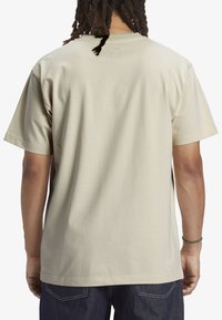 Beige bavlněné tričko s krátkým rukávem, kulatým výstřihem a malou etiketou na zadní straně. Hladká textura s volným střihem.
