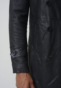 Zwarte leren jas met een dubbelbreasted ontwerp, grote knopen en een knoopmanchetaccent met gestructureerde mouwdetails.