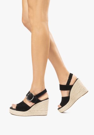 Sandales à talons compensés en suede noir avec une base enroulée de corde, bout ouvert, bride de cheville, et un accent en métal argenté bien visible.