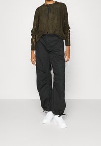 Olivgrön, texturerad stickad tröja med slitna detaljer, ihop med lösfittande svarta cargobyxor och vita sneakers.