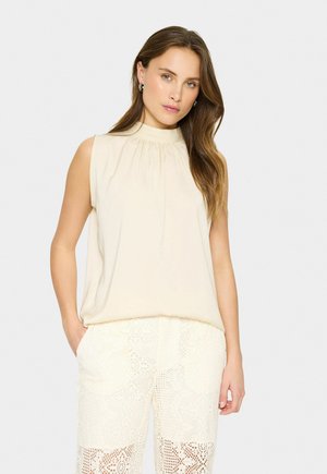 Vrouw met lang bruin haar draagt een mouwloze beige blouse en witte kanten broek, staand tegen een effen lichte achtergrond.