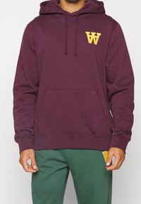 Vínová mikina s kapucí s přední kapsou, žlutým logem, nastavitelnou šňůrkou na kapuci a žebrovanými lemy. Doplněná zelenými tepláky.