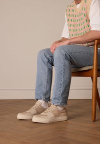 Beige Wildlederschuhe mit einer glatten Oberfläche, Schnürdesign und weißen Gummisohlen. Getragen mit hellblauen Jeans und weißen Socken, sitzend auf einem Holstuhl.