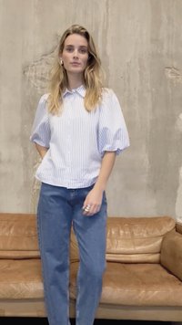 Gestreept blauw-wit overhemd met korte mouwen en knoopsluiting, met opgeblazen mouwen, gecombineerd met een high-waist blauwe jeans. Achtergrond: neutraalkleurige betonnen muur.