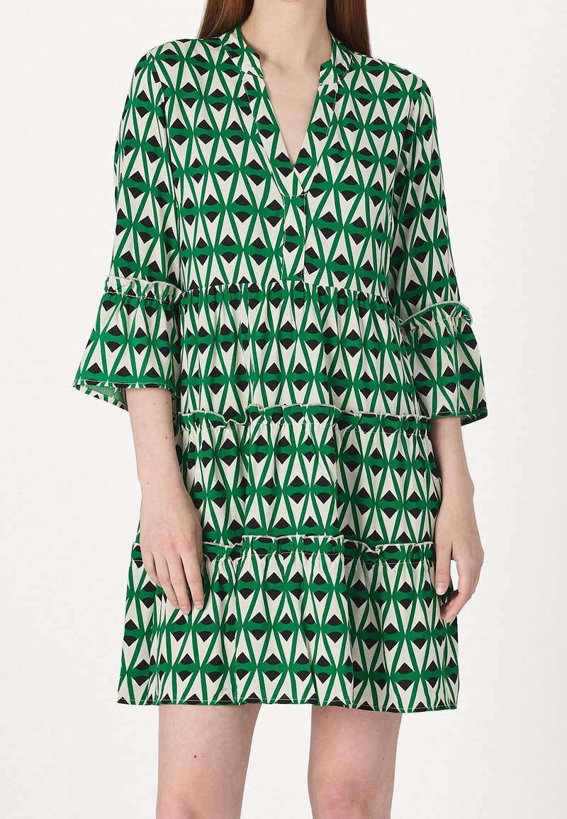 Femme portant une robe à motif géométrique vert et blanc avec des manches trois-quarts à volants et une jupe à volants superposés.