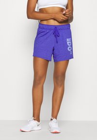 Osoba v purpurových sportovních šortkách s nápisem "JUST DO IT.", bílých teniskách a bílém crop topu, stojící na jednobarevném pozadí.