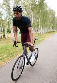 Un cycliste en maillot sombre et en shorts bordeaux roule sur un vélo de route gris sur un chemin pavé, entouré d'herbe verte et d'arbres blancs.
