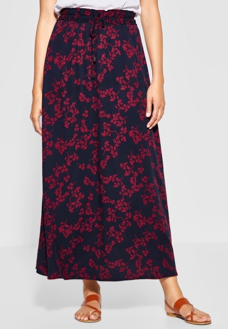 Femme portant une jupe longue bleu marine à motifs floraux rouges, un haut blanc et des sandales plates marron, debout devant un fond uni.