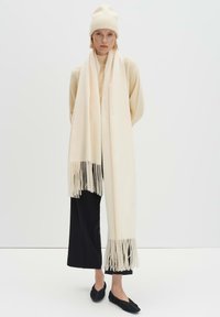 Lange crèmekleurige sjaal met franjes aan de uiteinden, gemaakt van zachte stof, gedragen over een beige coltrui en zwarte broek met wijde pijpen, gecombineerd met een bijpassende muts.