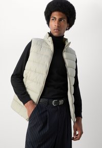 Young man with afro hairstyle wearing beige quilted vest over black turtleneck and pinstripe trousers with black belt, looking right.