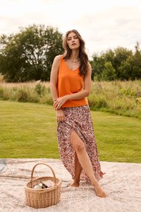 Femme portant un haut orange sans manches et une jupe à motifs, debout pieds nus sur une couverture à côté d'un panier de pique-nique en osier dans un champ herbeux.