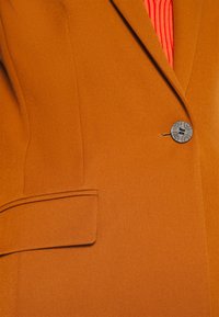 Gros plan sur un blazer orange brûlé avec un seul bouton sombre et une poche à rabat, porté sur un haut rouge texturé.