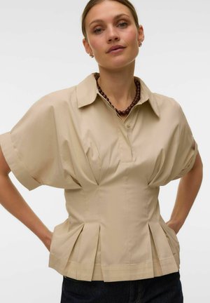 Kvinde iført beige bluse med korte ærmer med folder og en brun perlehalskæde, står med hænderne på hoften mod en hvid baggrund.